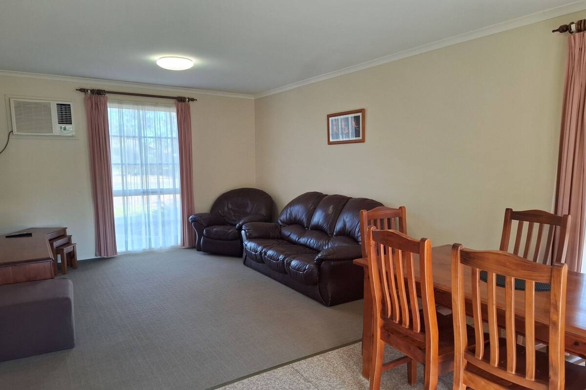 A living room with a leather sofa, wooden dining table, and chairs. There is a window with curtains, a picture on the wall, and an air conditioning unit on the wall.