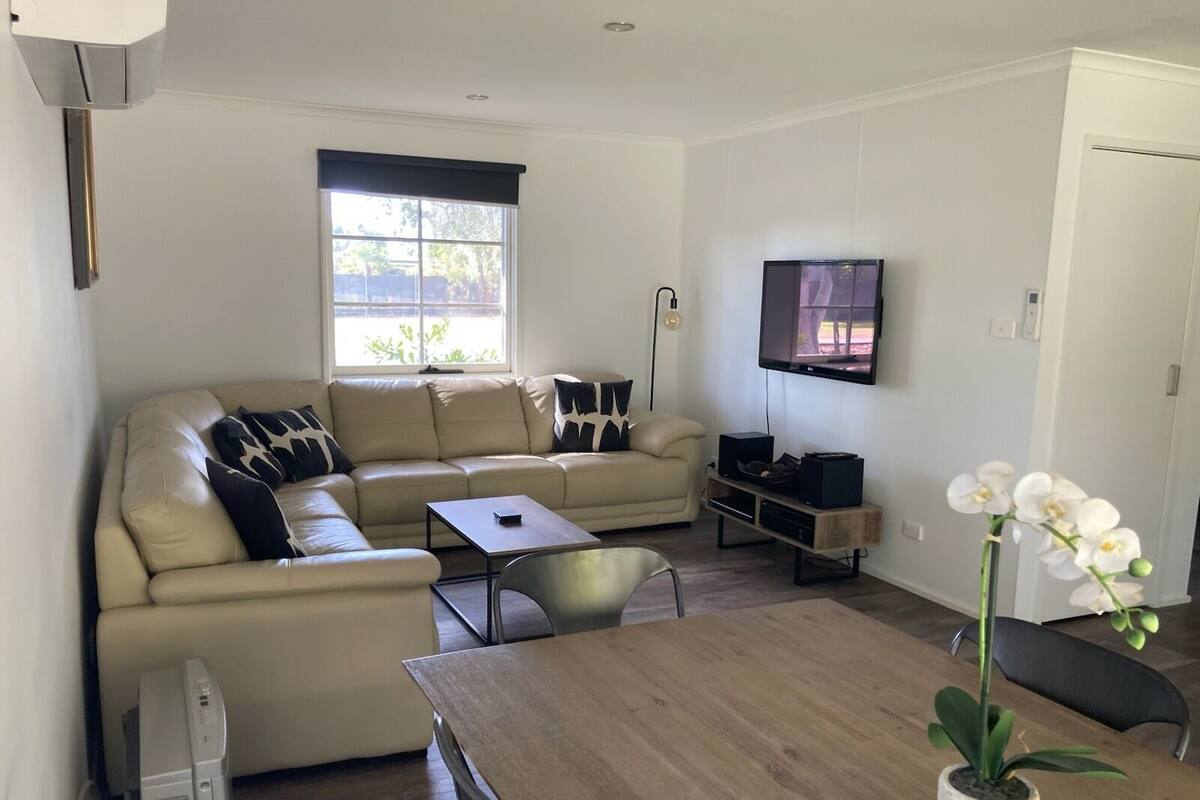 A modern living room with a beige sofa, a wooden coffee table, a flat-screen TV mounted on the wall, and a potted orchid on the table.