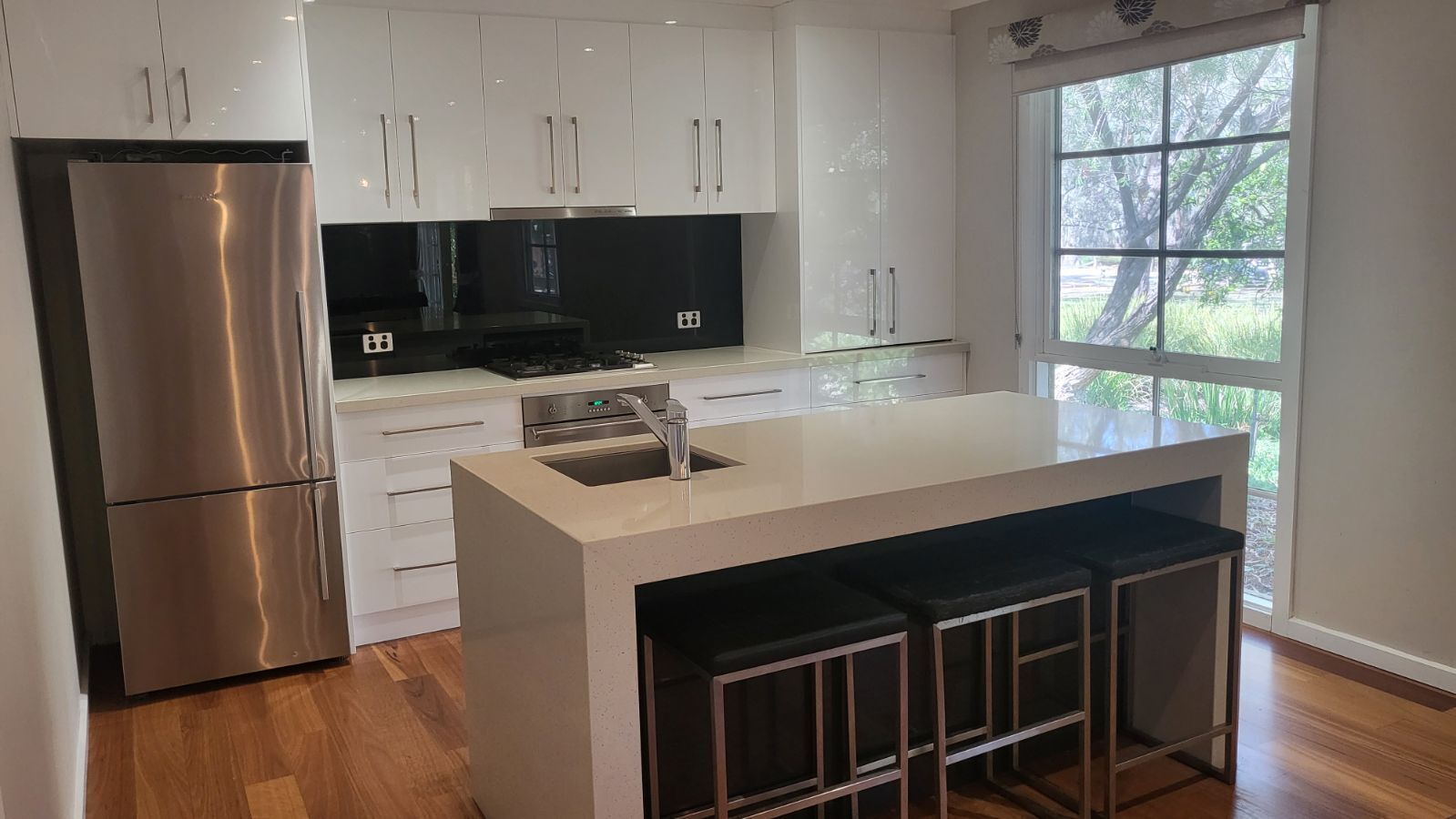 A modern kitchen with white cabinets, a stainless steel refrigerator, a black stove, and a large window with a view of trees.