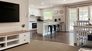 Una cocina moderna con armarios blancos, una isla central con taburetes, un televisor montado en la pared y un comedor con mesa y sillas.