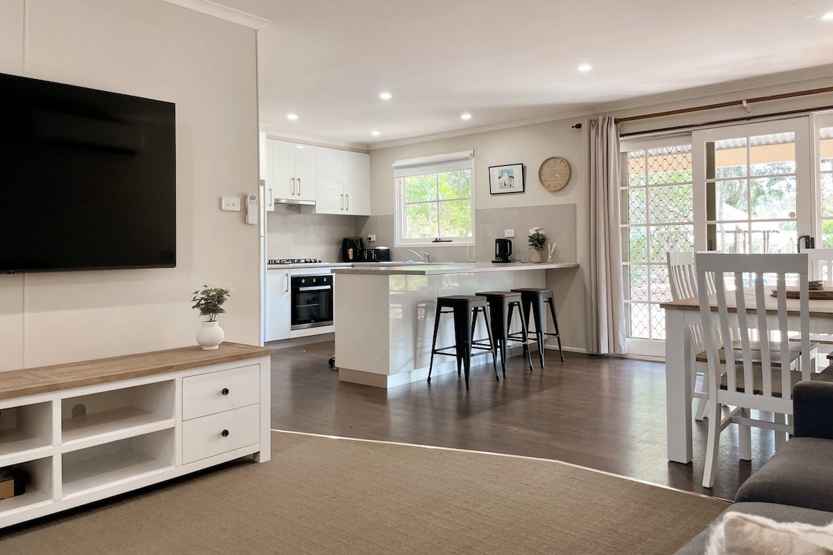 A modern kitchen with white cabinets, a central island with stools, a TV mounted on the wall, and a dining area with a table and chairs.