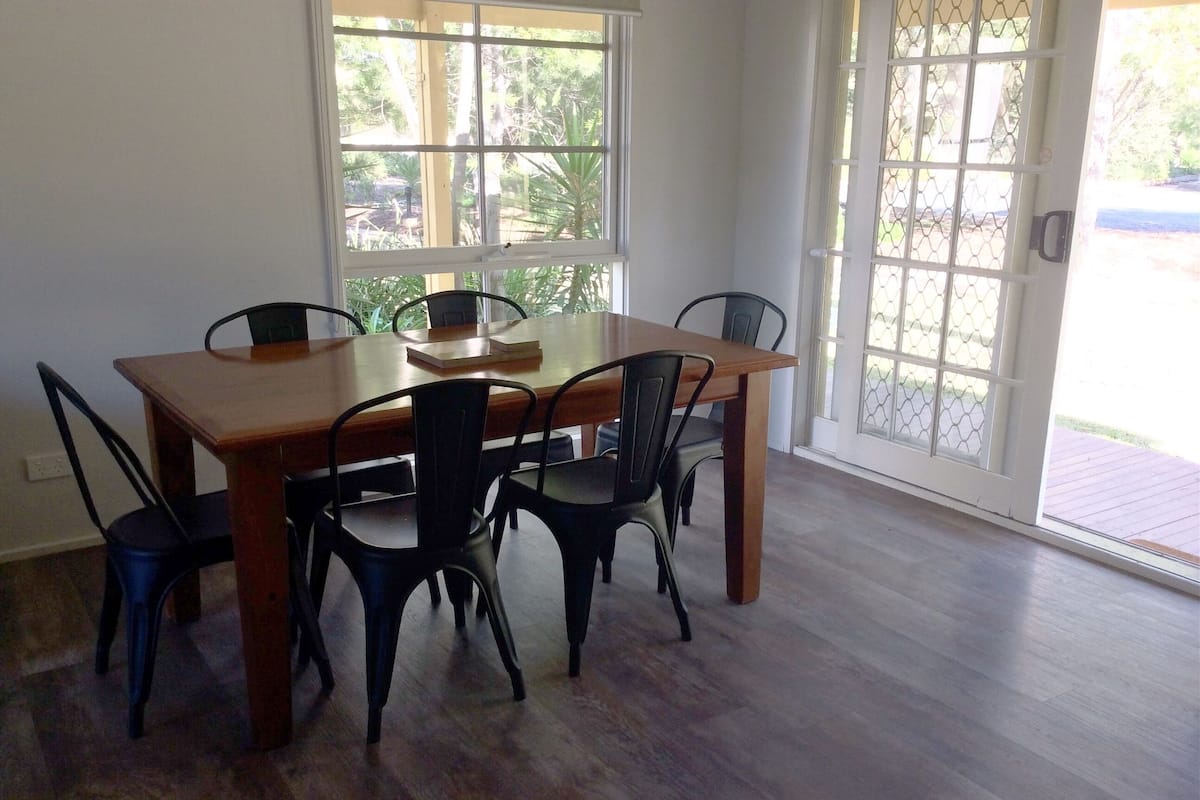 A dining area with a wooden table and six chairs, large windows, and a view of greenery outside.