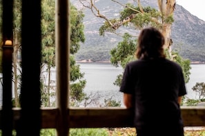 A person standing by a window overlooking a lake with trees and mountains in the background. - Freycinet Cottage 2 (Coles Bay)