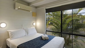 A hotel room with a bed, a window with a view of palm trees, an air conditioning unit, and a wall-mounted light fixture. - Discovery Parks - Ballina (East Ballina)