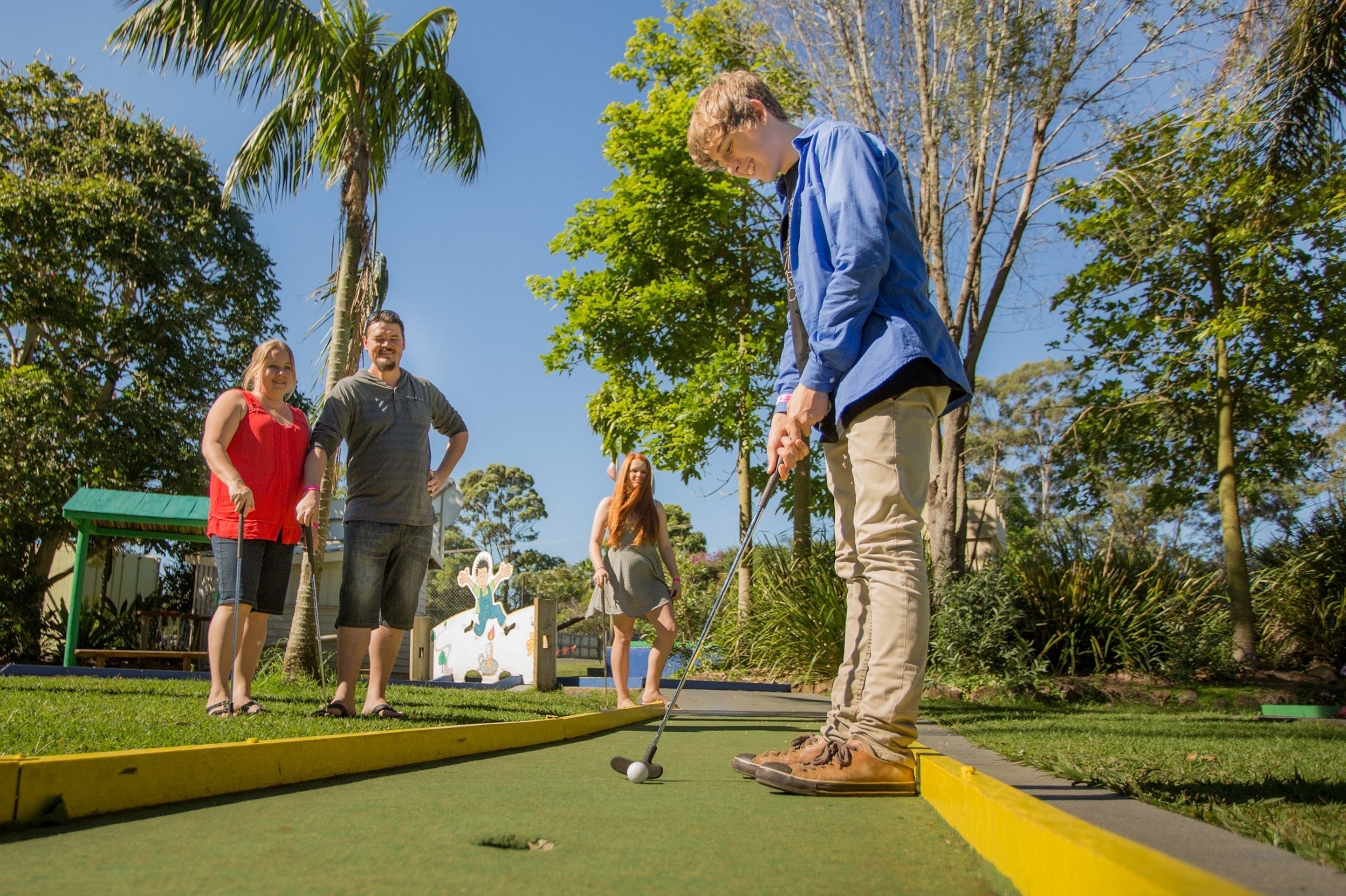 Un grupo de personas jugando croquet en un césped cuidado, con cielo despejado y palmeras al fondo.