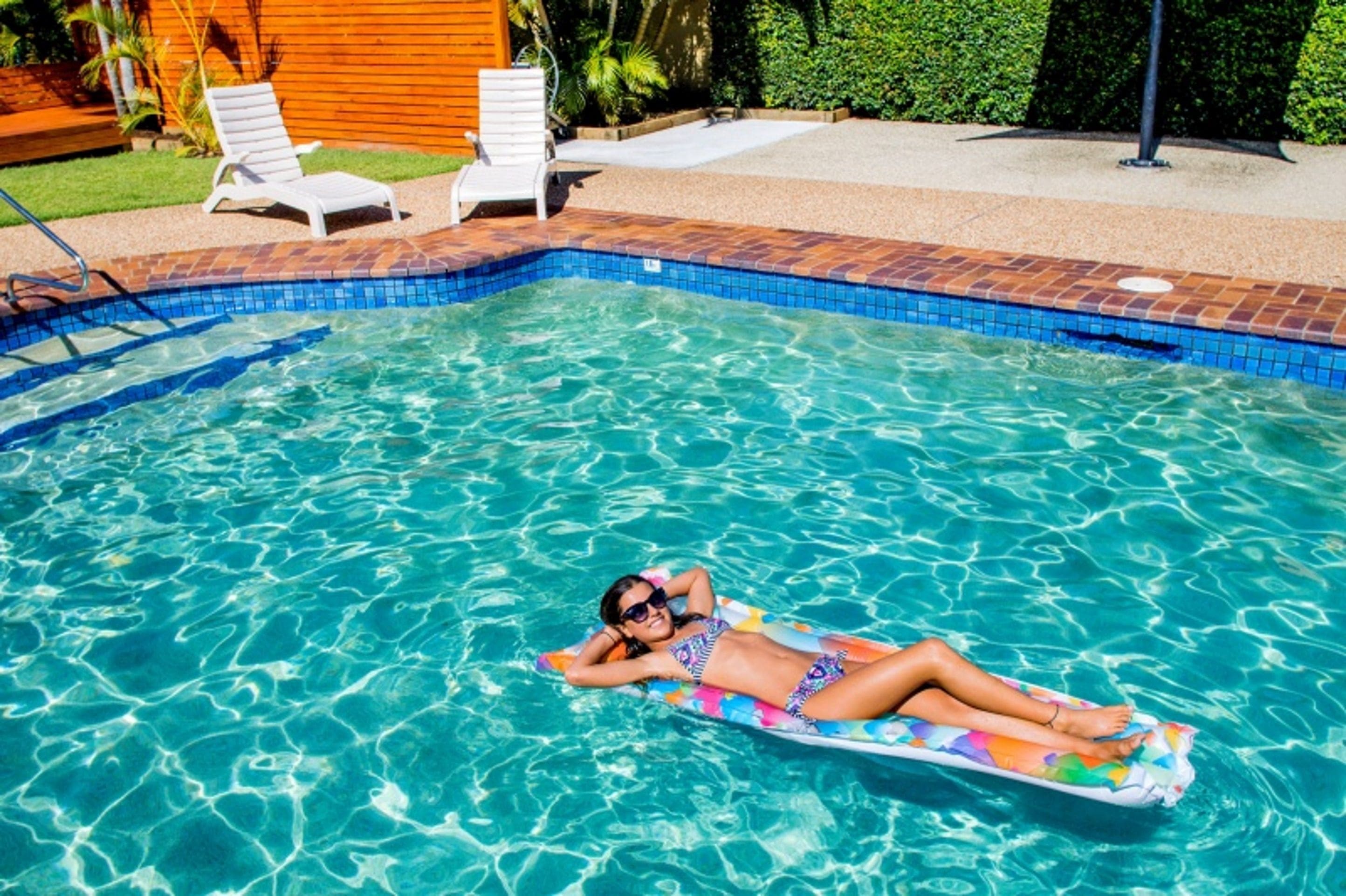 A swimming pool with clear blue water, a person lying on a colorful float, and two white lounge chairs in the background.