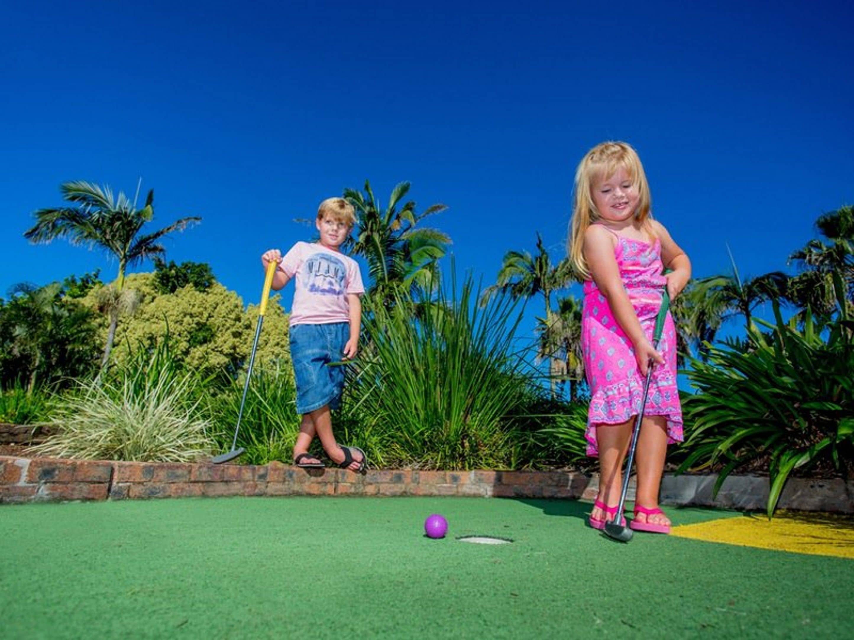 Dos niños jugando a croquet en un campo verde con palmeras al fondo.