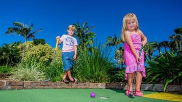 Dos niños jugando a croquet en un campo verde con palmeras al fondo.