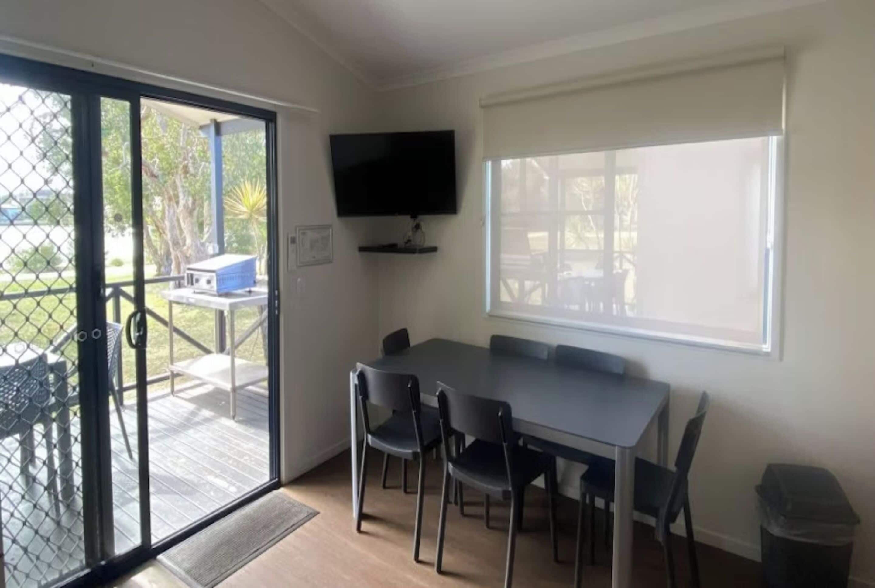 Habitación con una mesa de comedor, cuatro sillas, un televisor montado en la pared y un ventanal corredizo que da a un espacio exterior con mesa y sillas.
