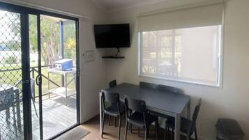 Habitación con una mesa de comedor, cuatro sillas, un televisor montado en la pared y un ventanal corredizo que da a un espacio exterior con mesa y sillas.