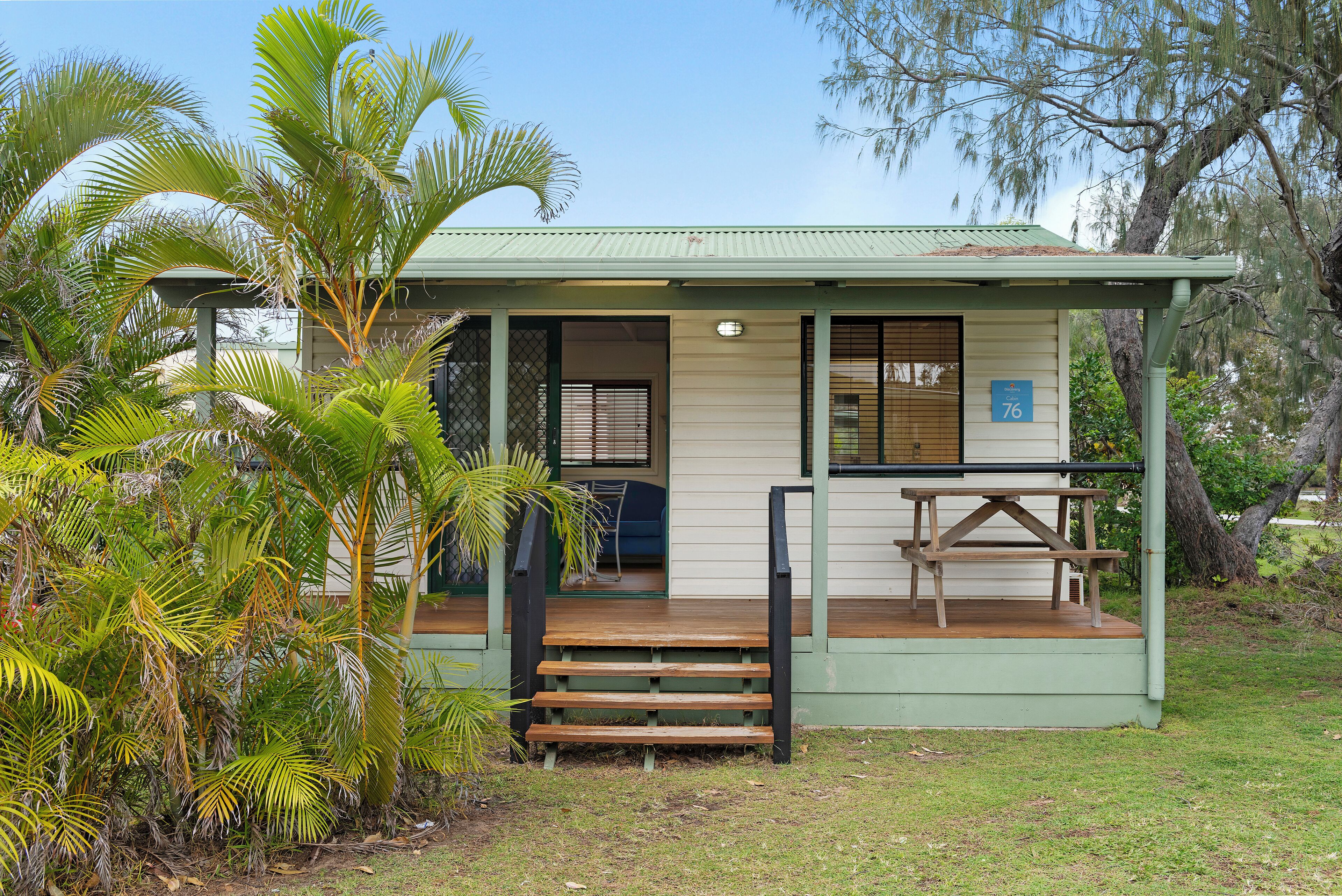 A small, single-story building with a green roof and a wooden deck. There is a bench and a table on the deck.
