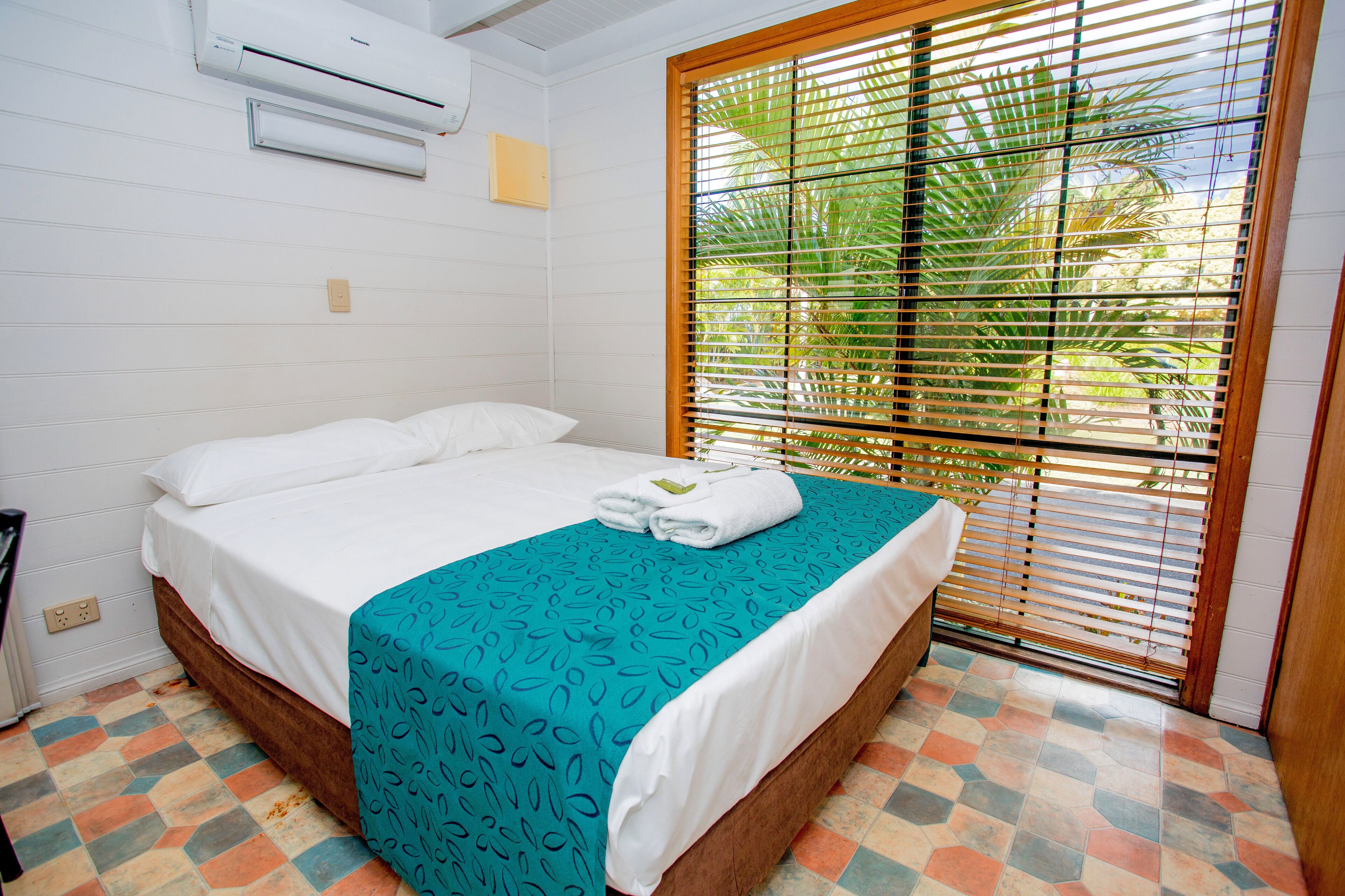 A hotel room with a bed, air conditioning unit, window with blinds, and a tiled floor.