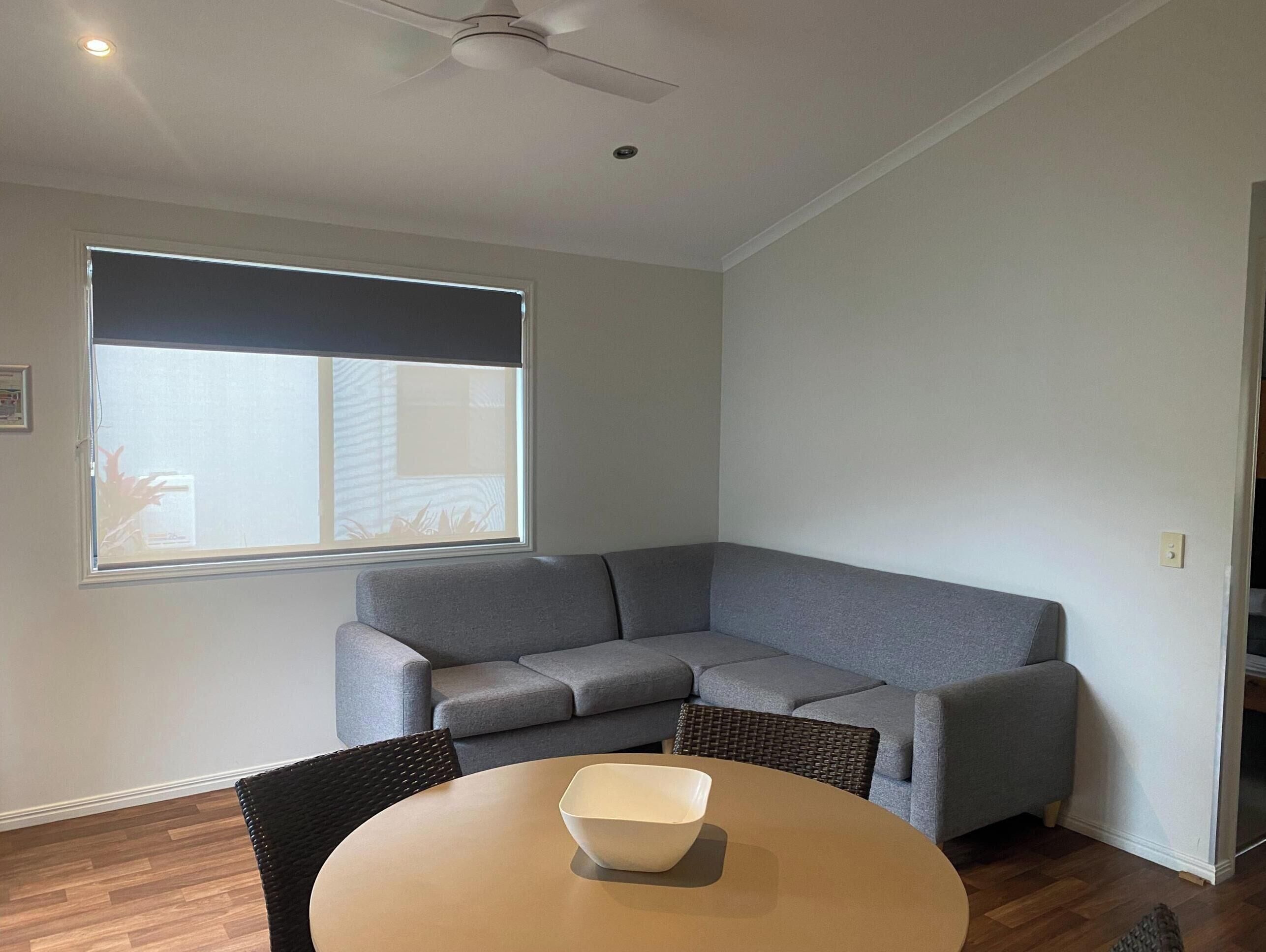 Una sala de estar moderna con un sofá seccional gris, una mesa redonda con un cuenco, un ventilador de techo y un ventanal con persianas.