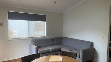 Una sala de estar moderna con un sofá seccional gris, una mesa redonda con un cuenco, un ventilador de techo y un ventanal con persianas.