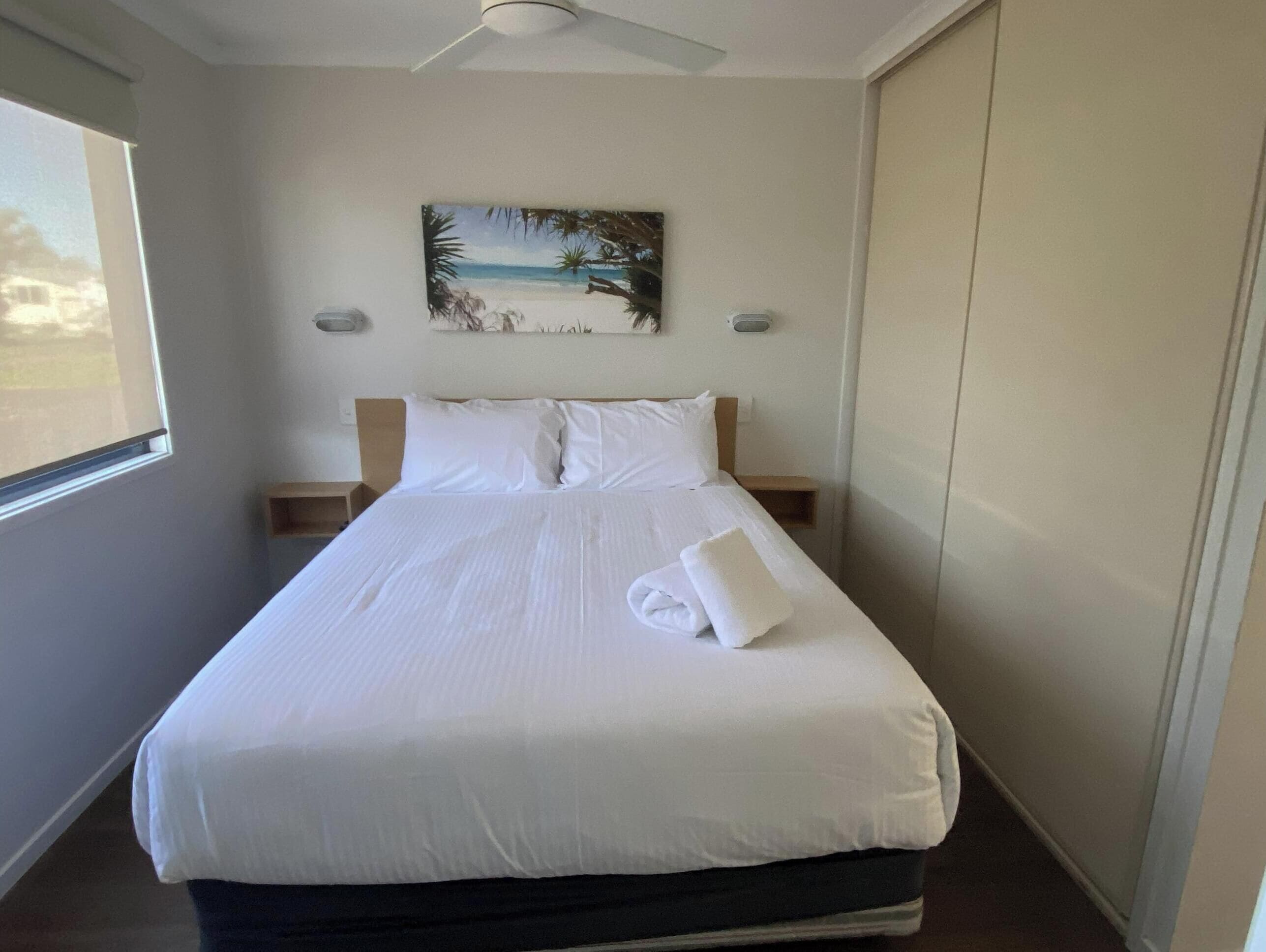 Una cama bien hecha con sábanas blancas, un cuadro con una playa en la pared y un ventilador de techo.