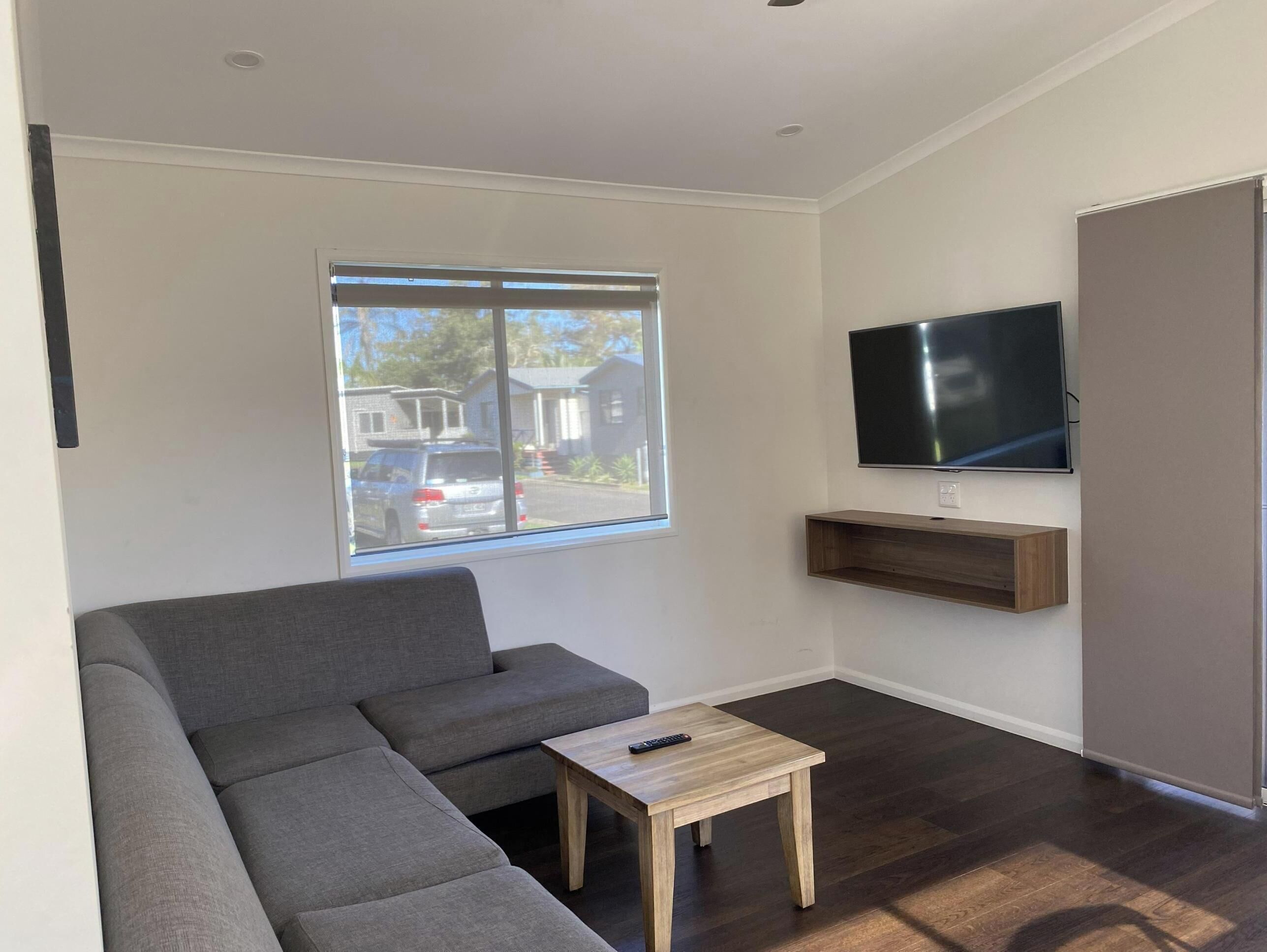 A living room with a grey sectional sofa, a small wooden coffee table, a mounted TV, and a window with a view of houses and a car.