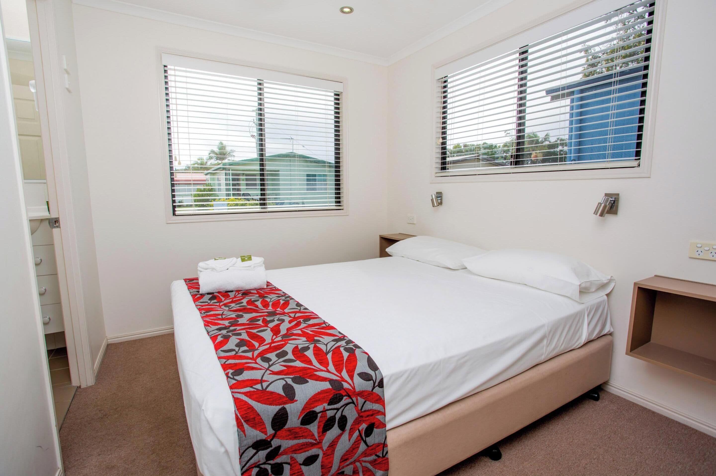 A bedroom with a bed, two windows with blinds, a bedside table, and a carpeted floor.