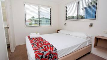 A bedroom with a bed, two windows with blinds, a bedside table, and a carpeted floor.