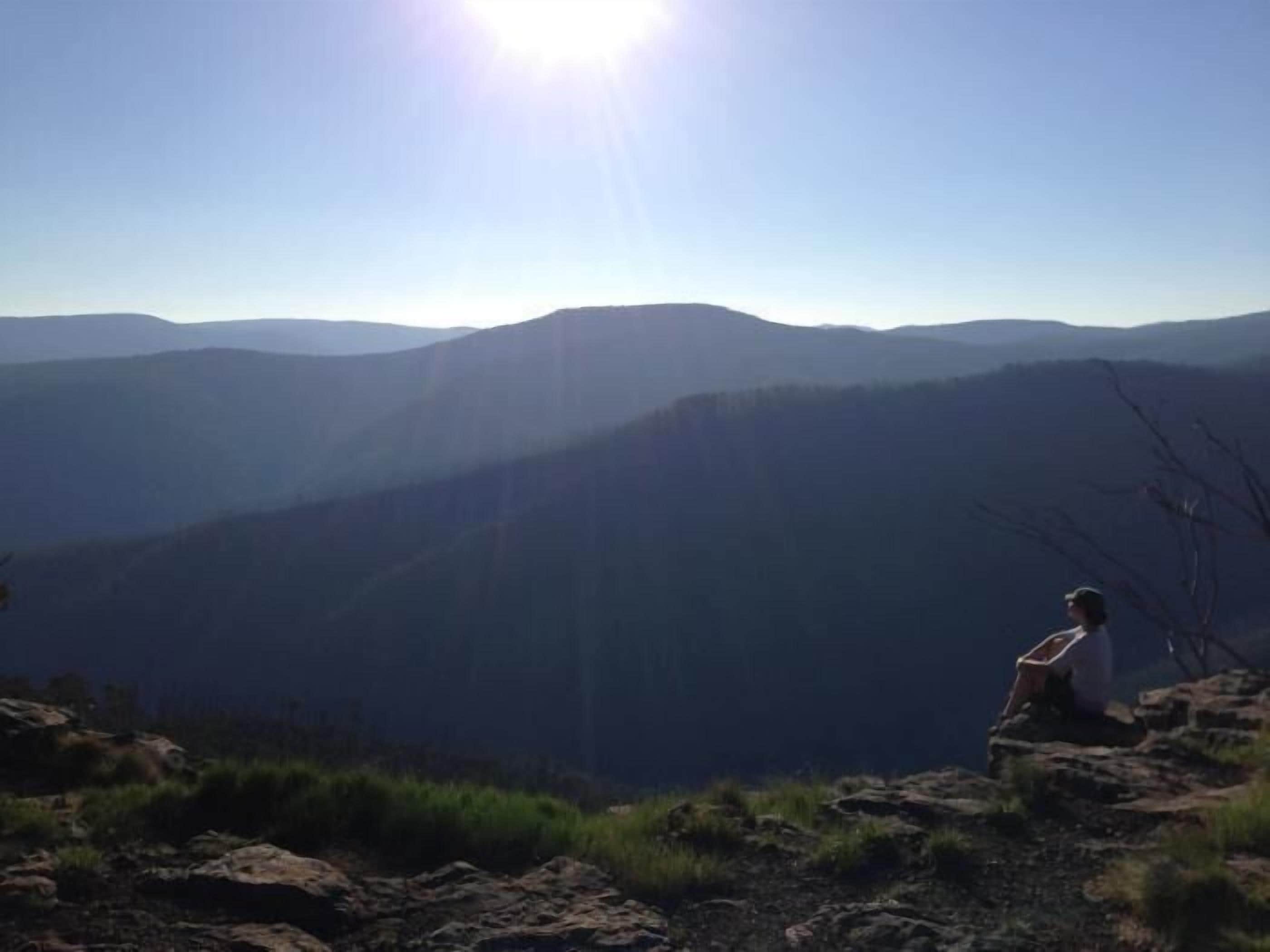 Una persona sentada sobre un afloramiento rocoso, contemplando una cadena montañosa con el sol brillando al fondo.
