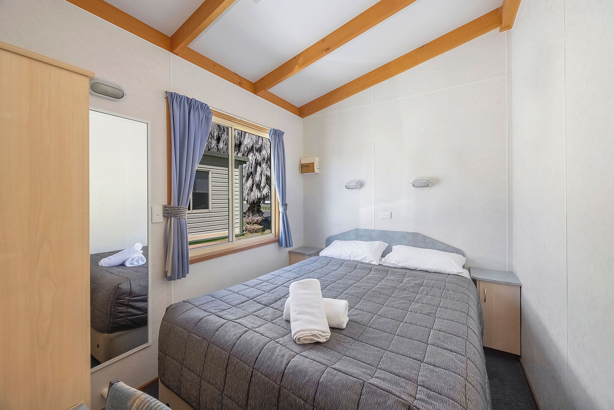 Un dormitorio con cama, mesita de noche, espejo y ventana con cortinas.