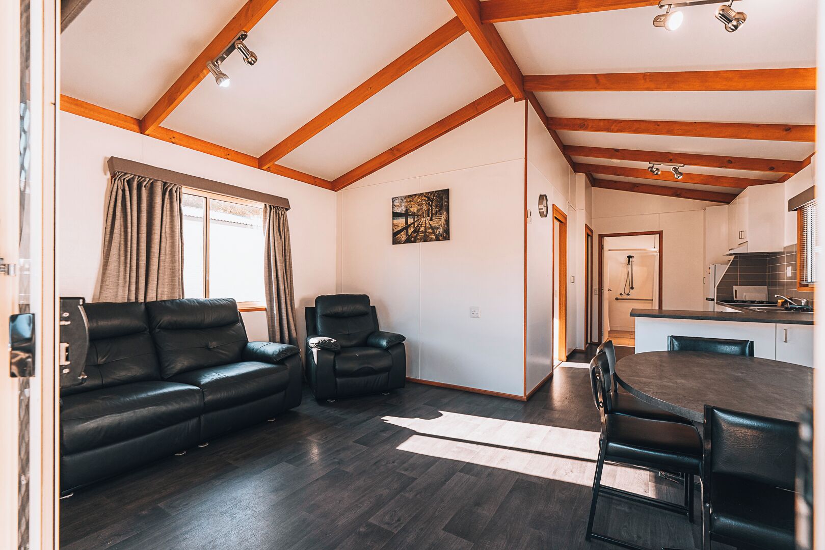 A modern living room with a dark wood floor, a black leather sofa, a recliner, and a dining table with chairs. A kitchen is visible in the background.