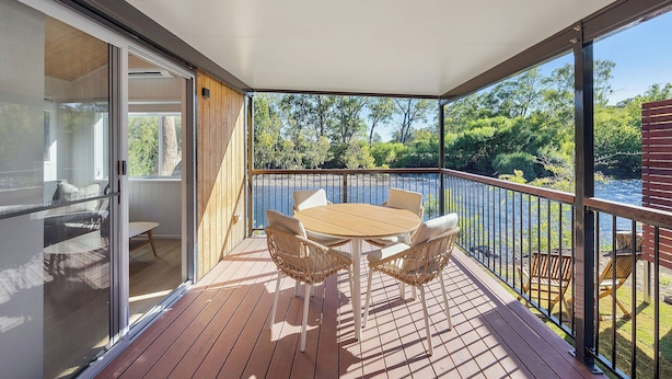 A wooden deck with a table and chairs, a glass door leading to a room, and a view of a lake and trees. - Reflections Tumut River (Tumut)