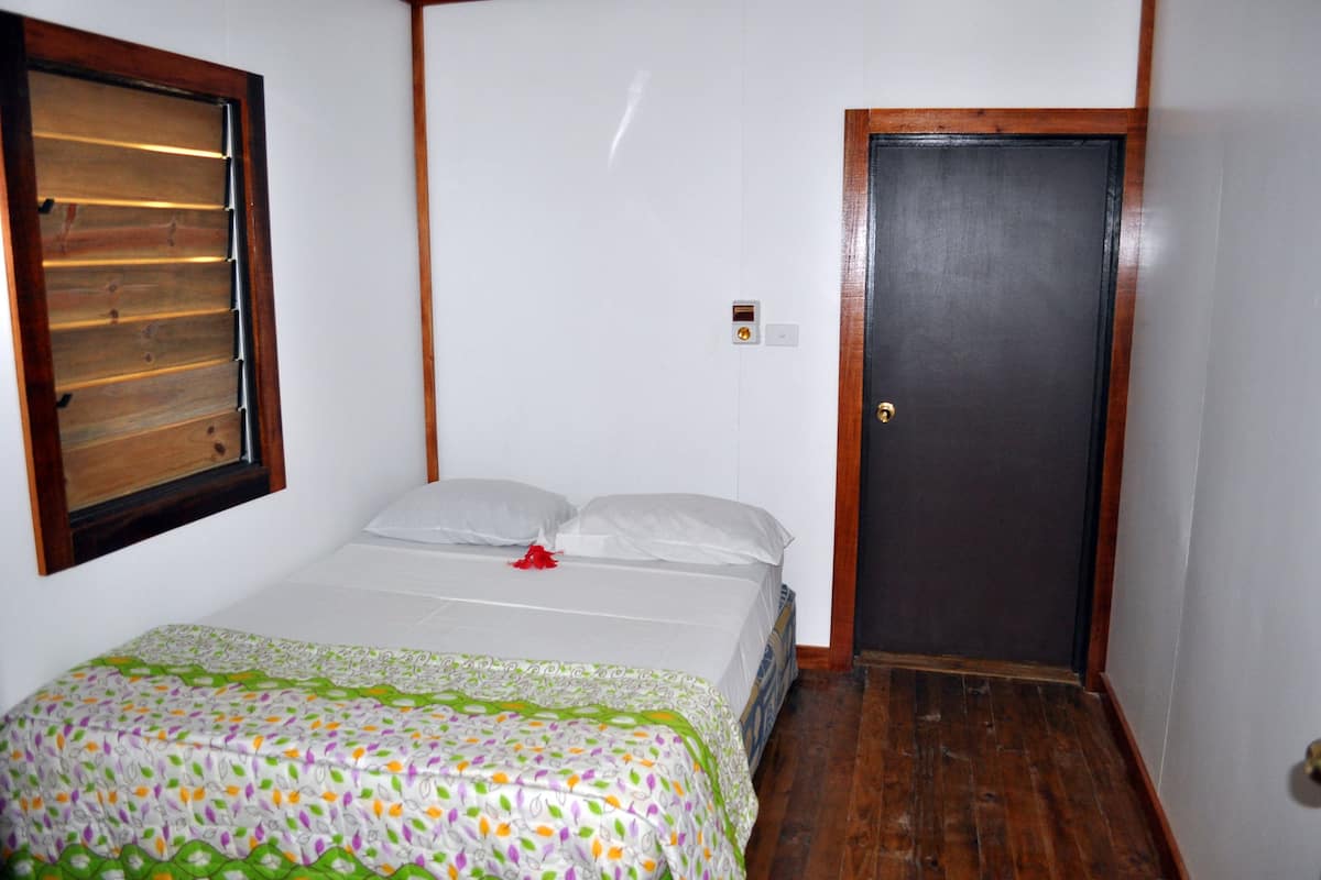 A small, single-bed room with a wooden door, a window with blinds, and a patterned bedspread.