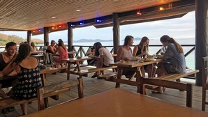 An outdoor dining area with wooden tables and benches, overlooking a body of water. - Ratu Kini Backpackers & Dive Resort (Mana Island)