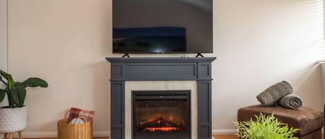 A living room with a fireplace, a flat-screen TV mounted above it, a brown sofa, and a potted plant.