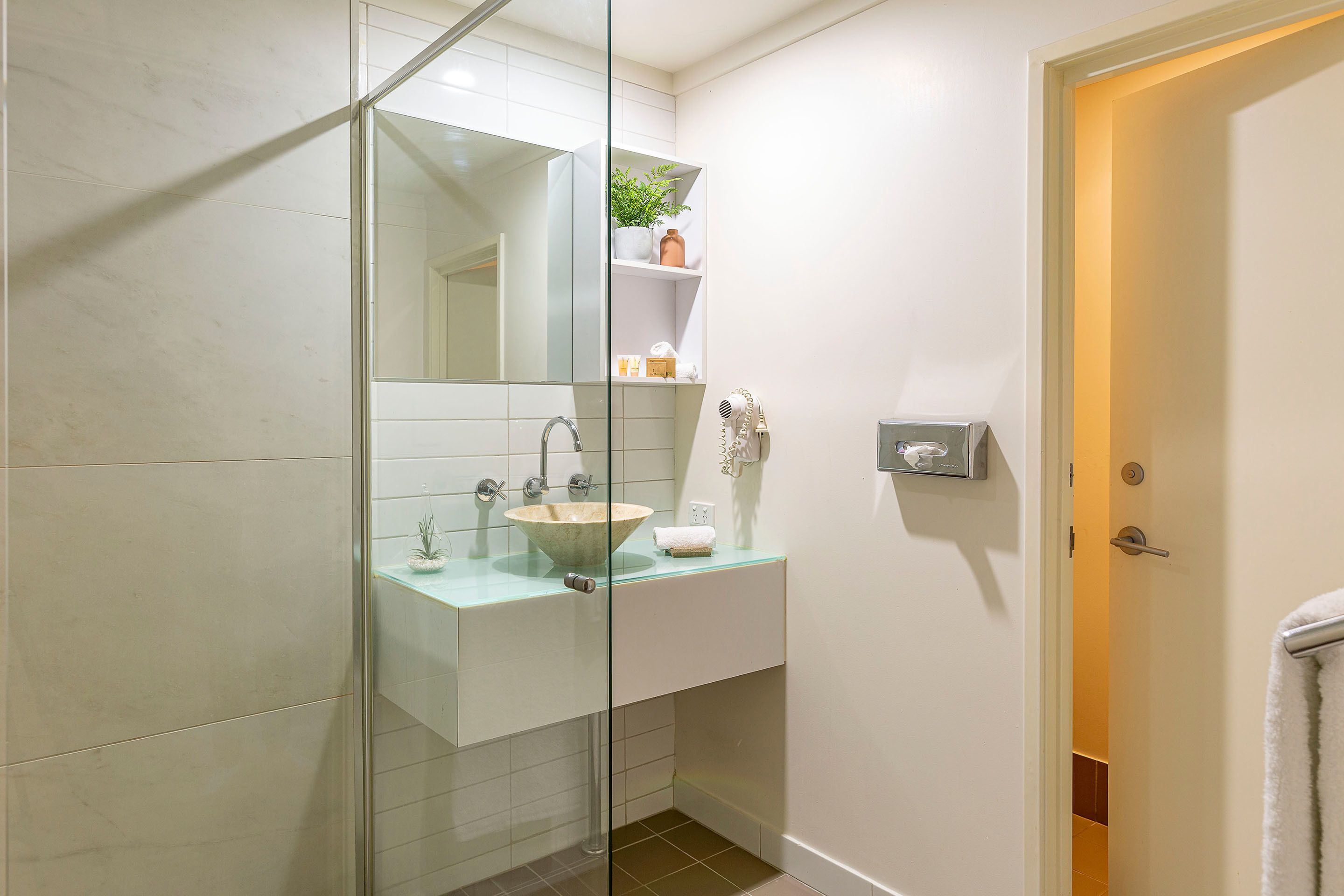 Un baño moderno con cabina de ducha de vidrio, un lavabo blanco con pileta y un estante pequeño con artículos de aseo.