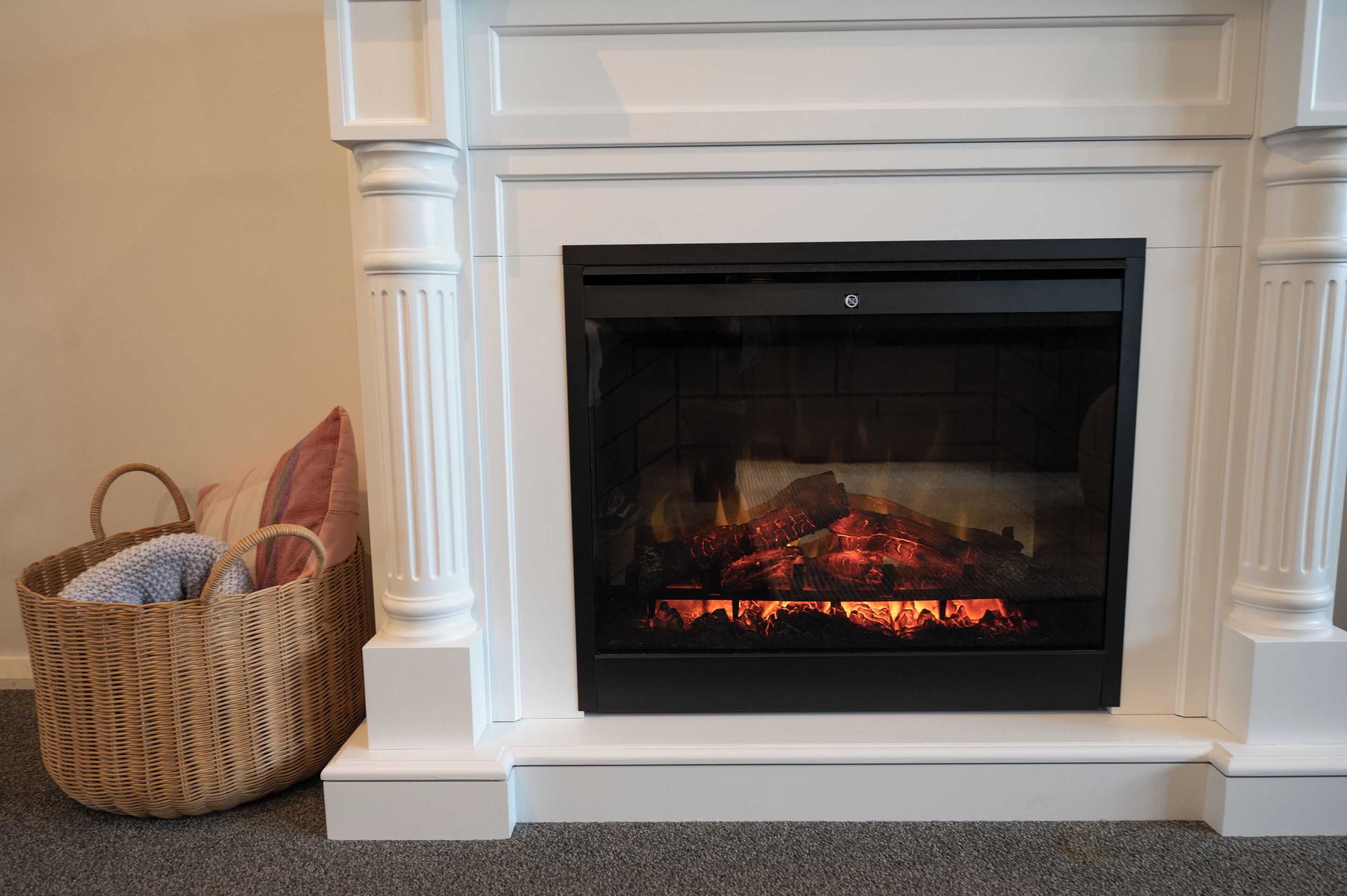 A fireplace with a fire, a wicker basket with cushions, and a white mantel.