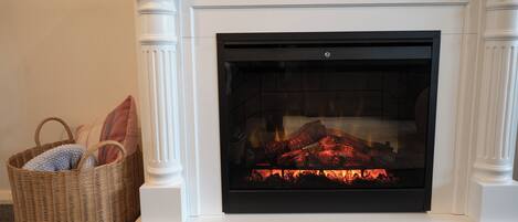 A fireplace with a fire, a wicker basket with cushions, and a white mantel.
