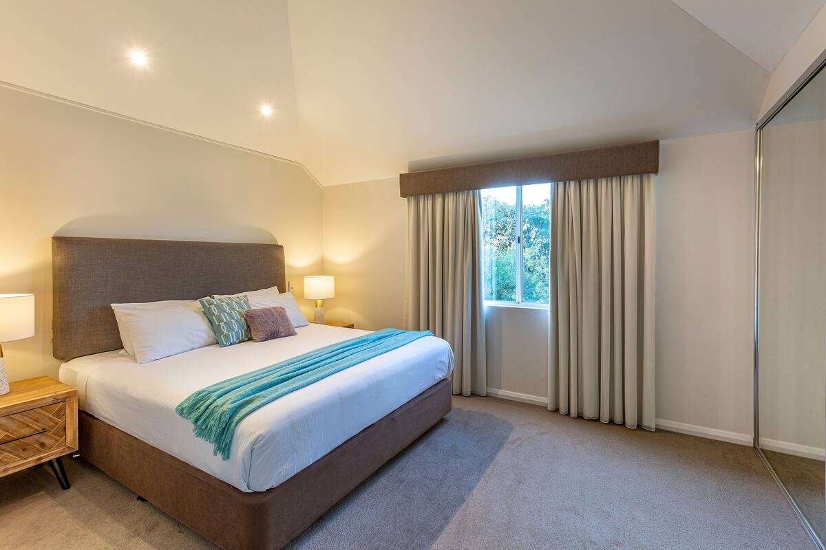 Habitación de hotel con una cama grande, una mesita de noche con lámpara, una ventana con cortinas y vistas a árboles.