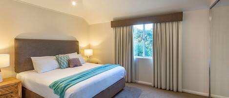 Habitación de hotel con una cama grande, una mesita de noche con lámpara, una ventana con cortinas y vistas a árboles.