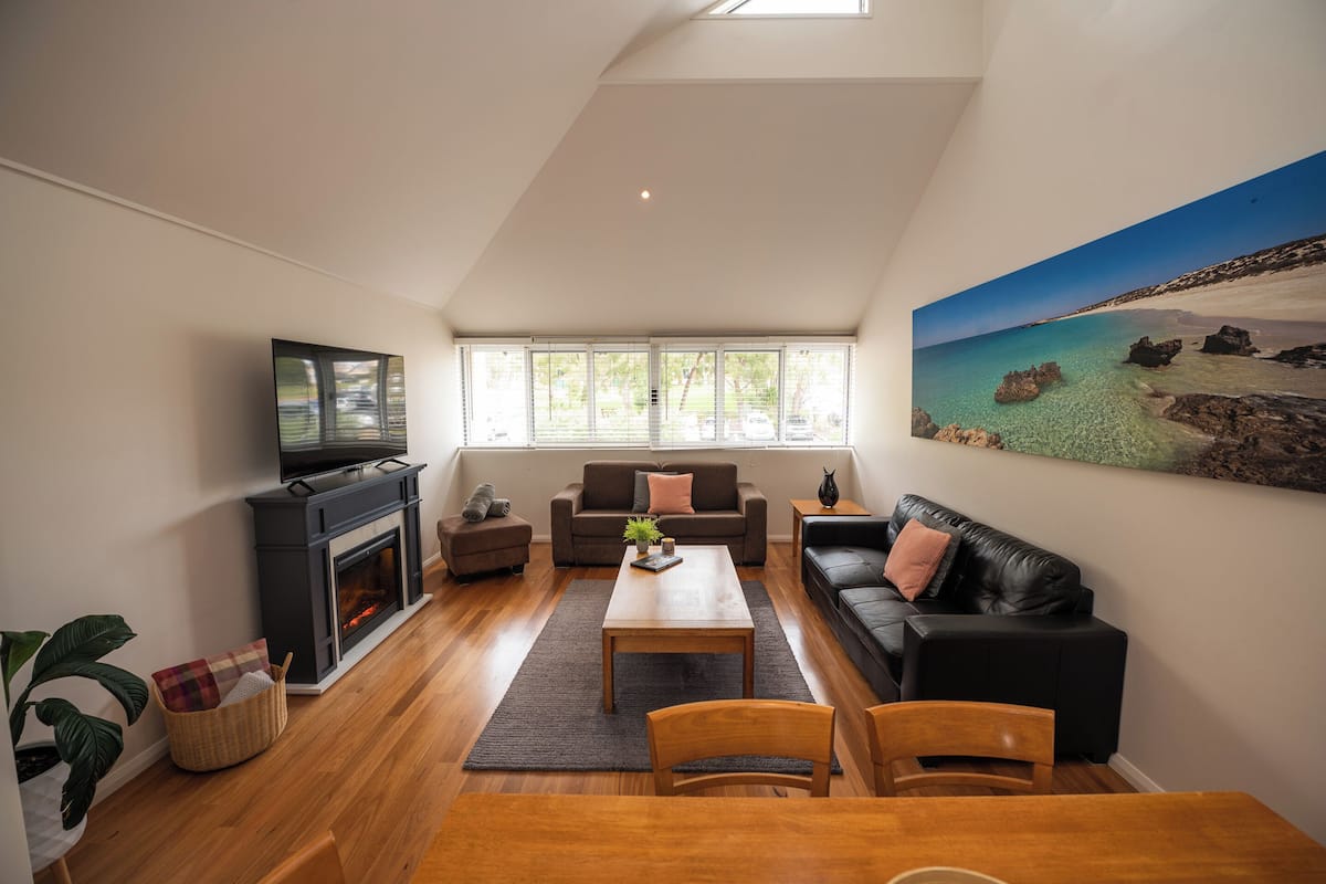 Una sala de estar con chimenea, televisor, sofá, mesa de centro y un cuadro con una escena de playa.