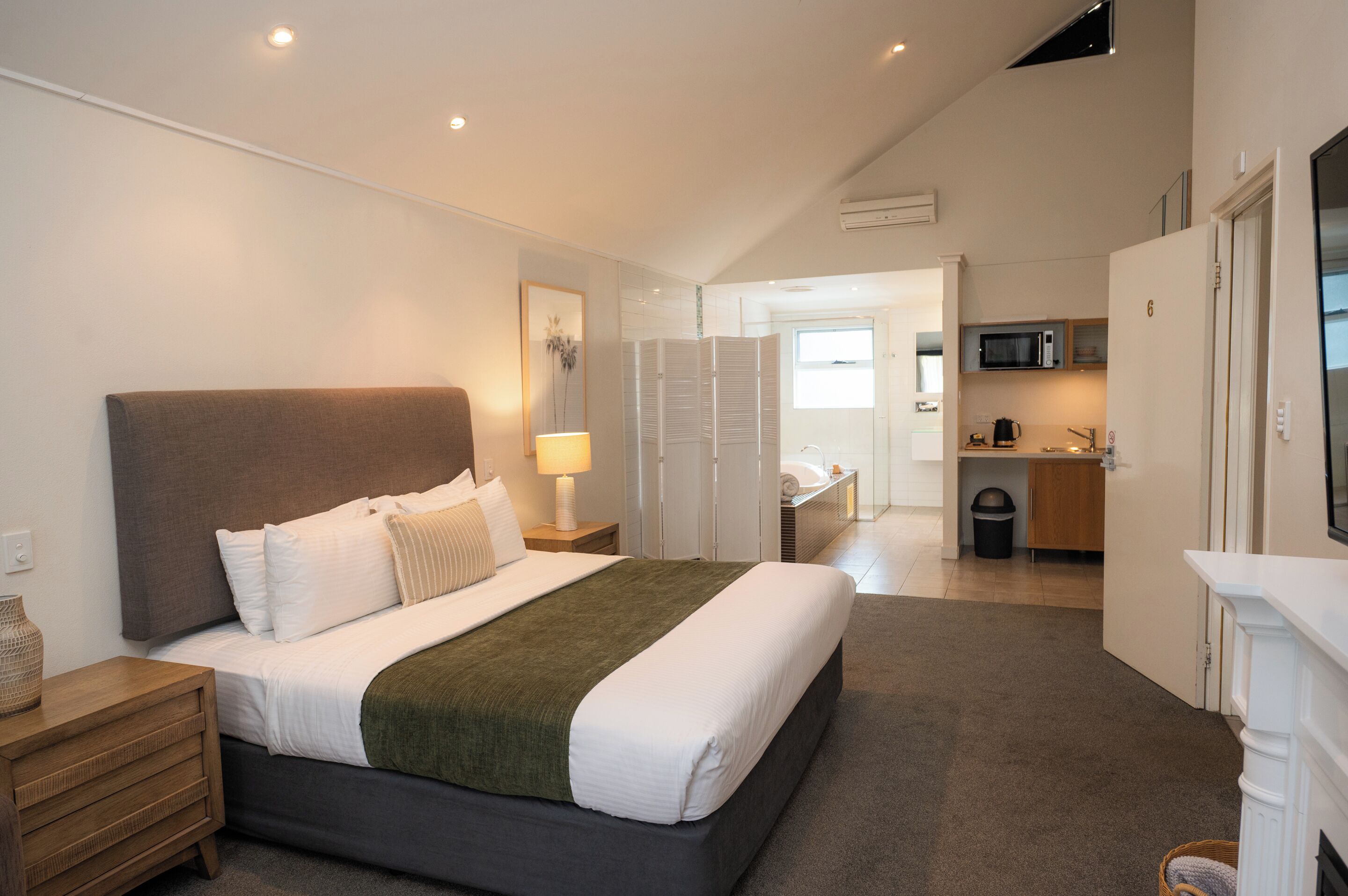 Habitación de hotel moderna con una cama grande, una mesita de noche, una lámpara y vistas a la cocina y el baño.