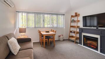 A living room with a sofa, a dining table with chairs, a TV, a fireplace, and a window with blinds.