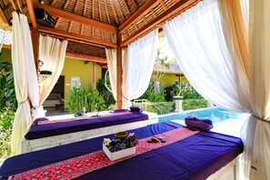 Un área de spa con dos camillas de masaje, cada una con una colcha morada y un tapete decorativo rojo.