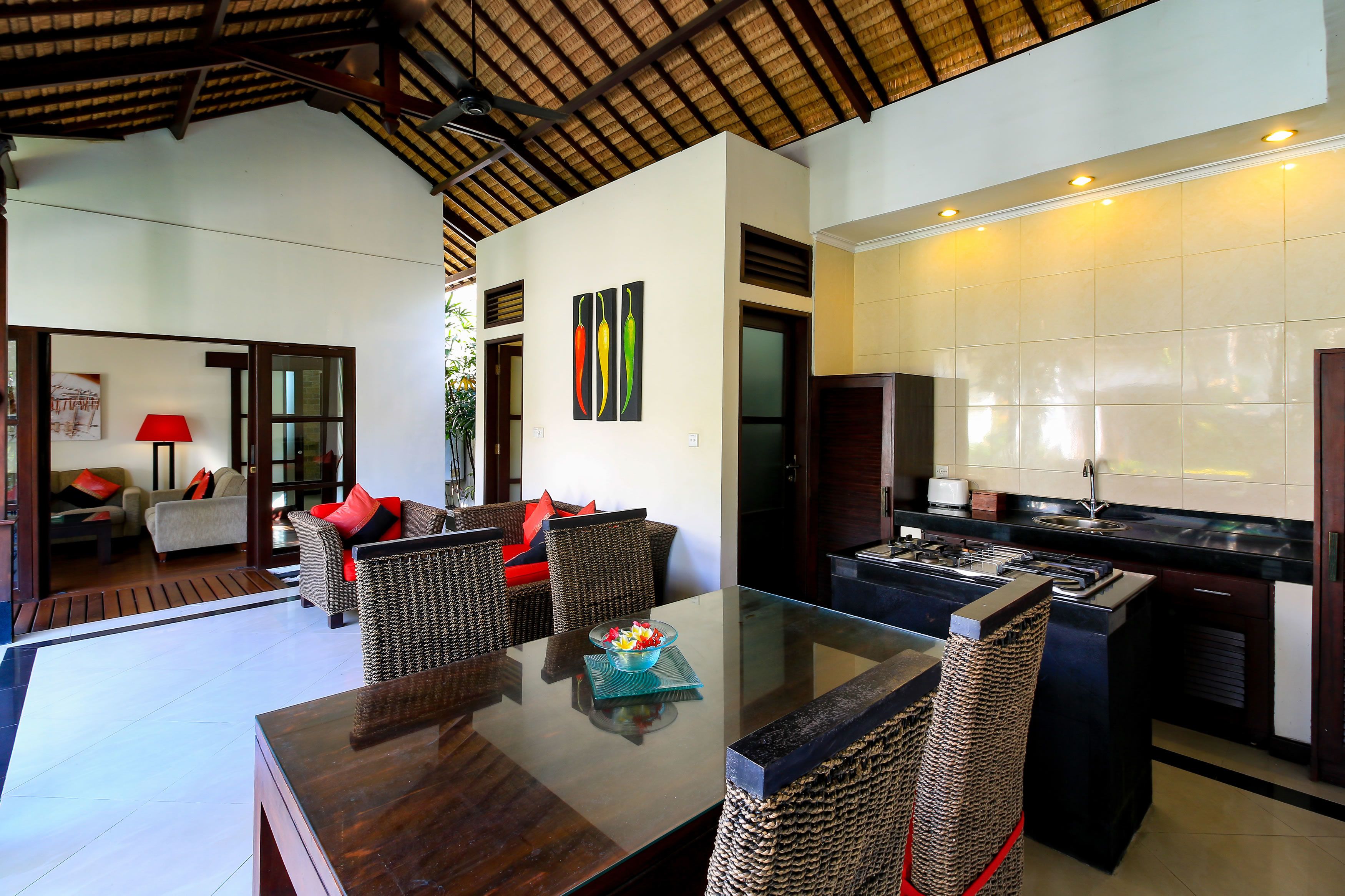 A modern living area with a glass-top dining table, wicker furniture, and a kitchen with a black countertop and white tiles.
