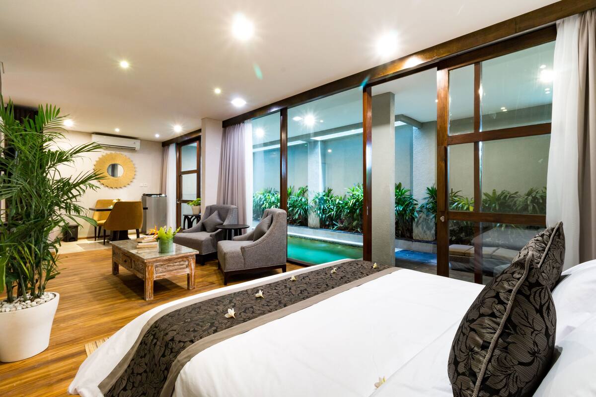 A hotel room with a large bed, a seating area with two armchairs and a coffee table, a small table with a lamp, a potted plant, and a view of a swimming pool through large glass doors.