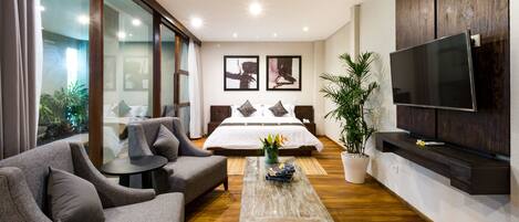 Habitación de hotel moderna con una cama grande, sofás grises, una mesa de centro, televisión y un ventanal con vistas a zonas verdes.