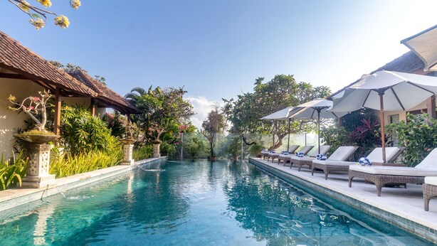 Une piscine entourée de chaises longues et de parasols, elle-même bordée d’une végétation luxuriante et d’un bâtiment de style traditionnel.