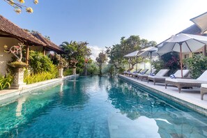 Una piscina con sillas de descanso y sombrillas, rodeada de exuberante vegetación y un edificio tradicional.