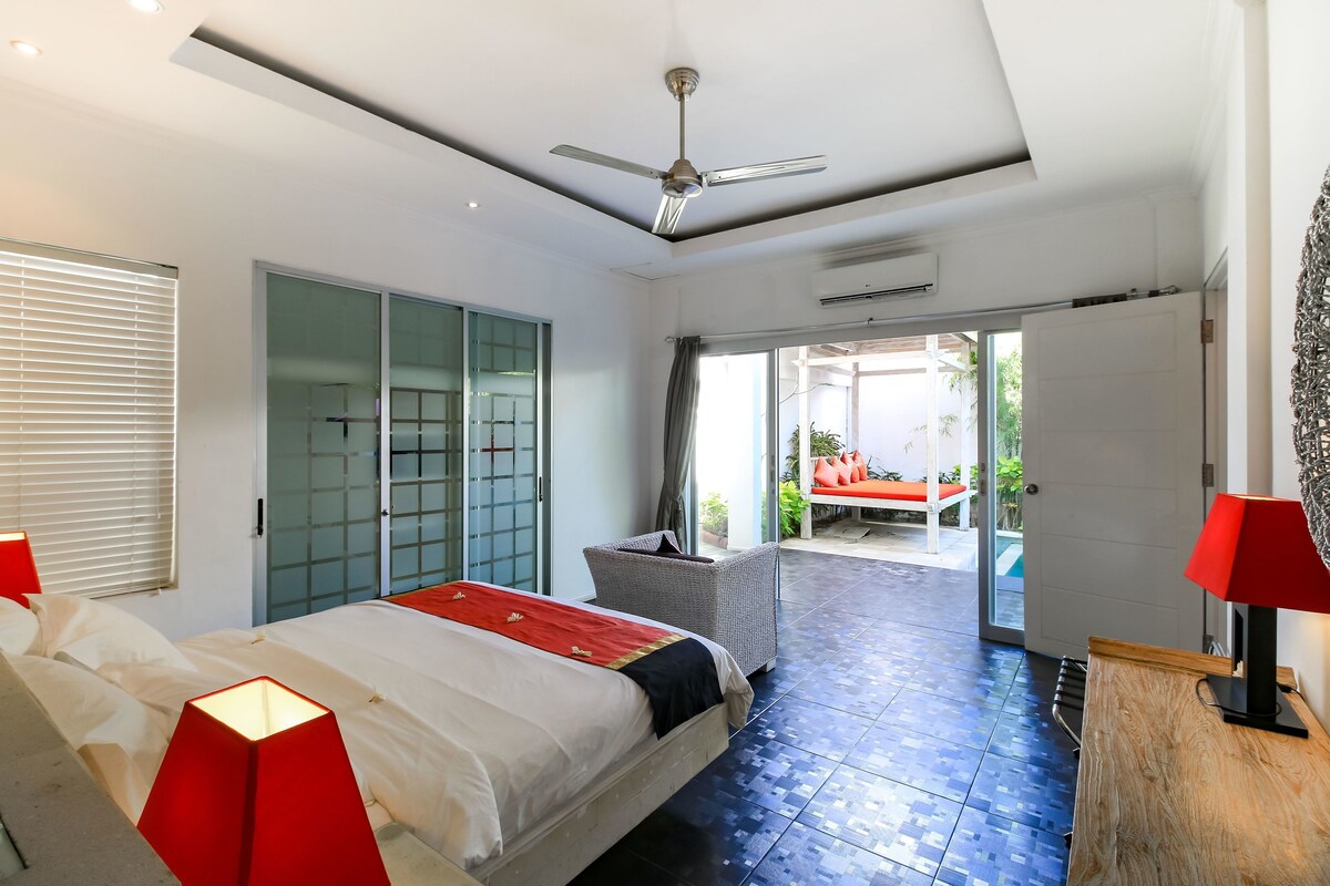 Una habitación de hotel moderna con una cama, un ventilador de techo, una lámpara roja y vistas a un área de estar al aire libre.