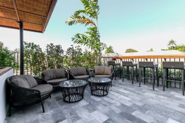 Un patio exterior con mobiliario de mimbre, mesas redondas y vistas a la vegetación y edificios circundantes.