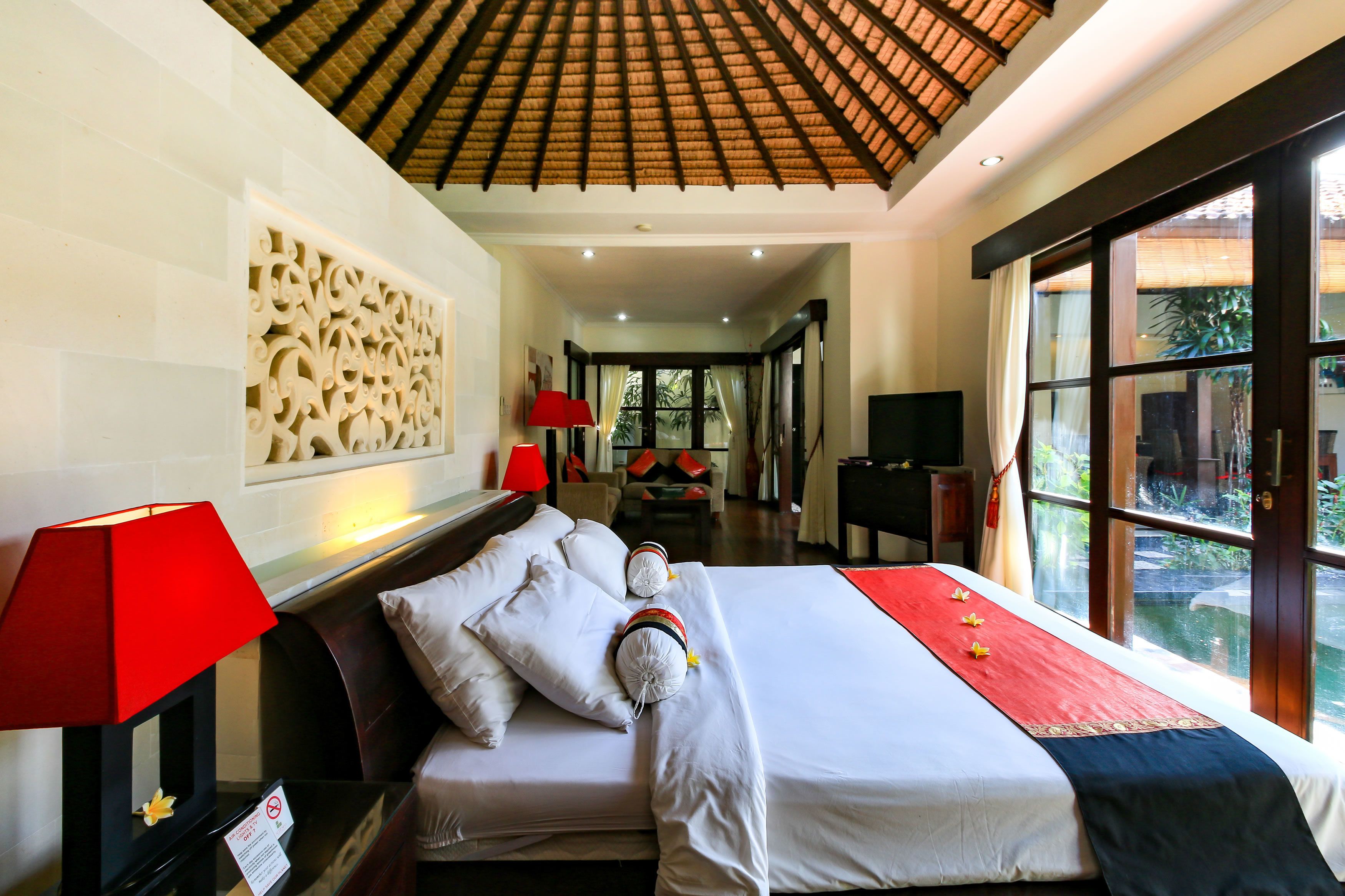 A hotel room with a bed, a red lamp, a decorative wall panel, and a view of greenery through the window.