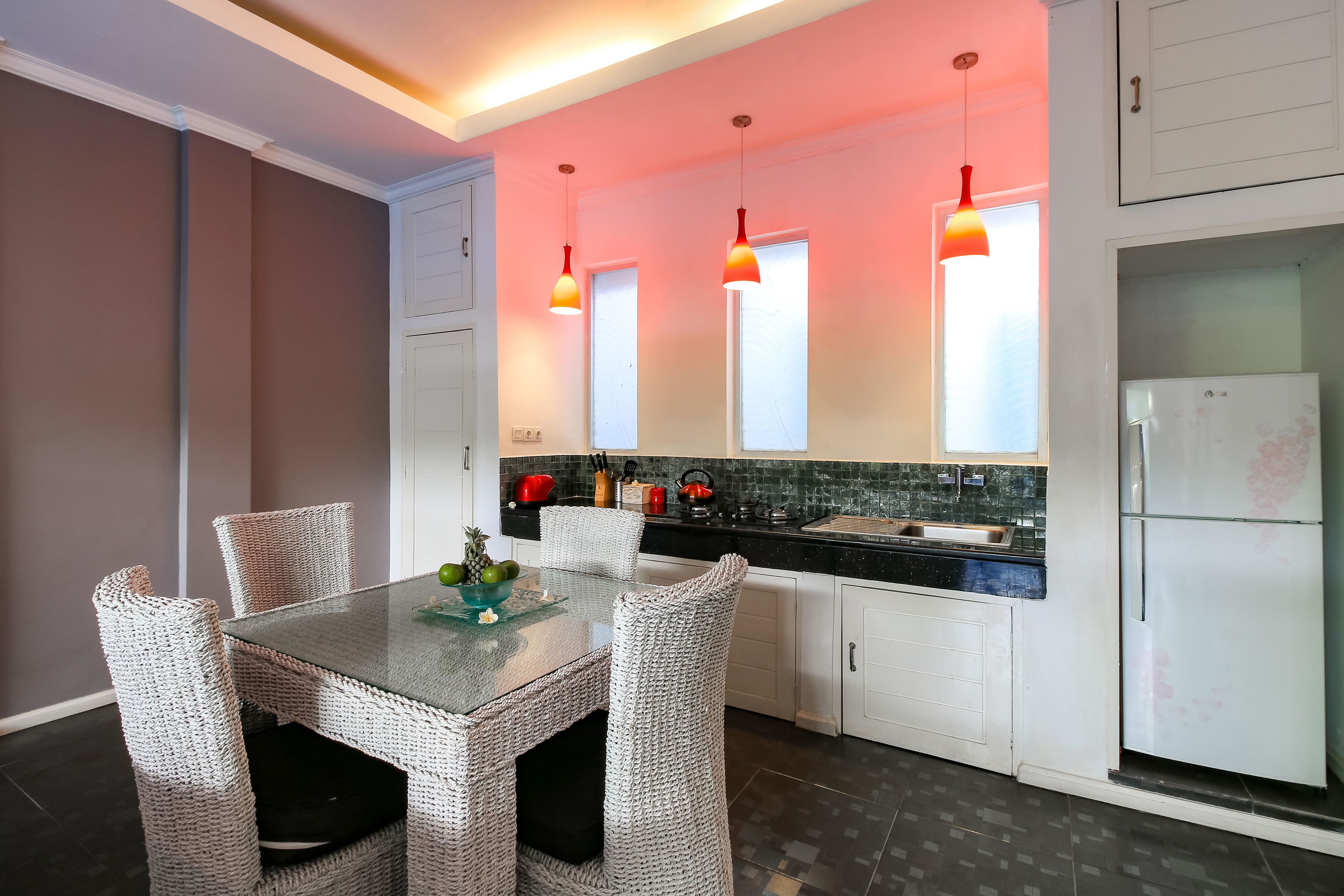 A modern kitchen with white cabinetry, a glass-top dining table, wicker chairs, and pendant lights.