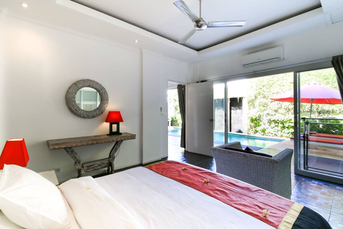 A bedroom with a bed, a nightstand with a lamp, a round mirror, and a view of an outdoor swimming pool.