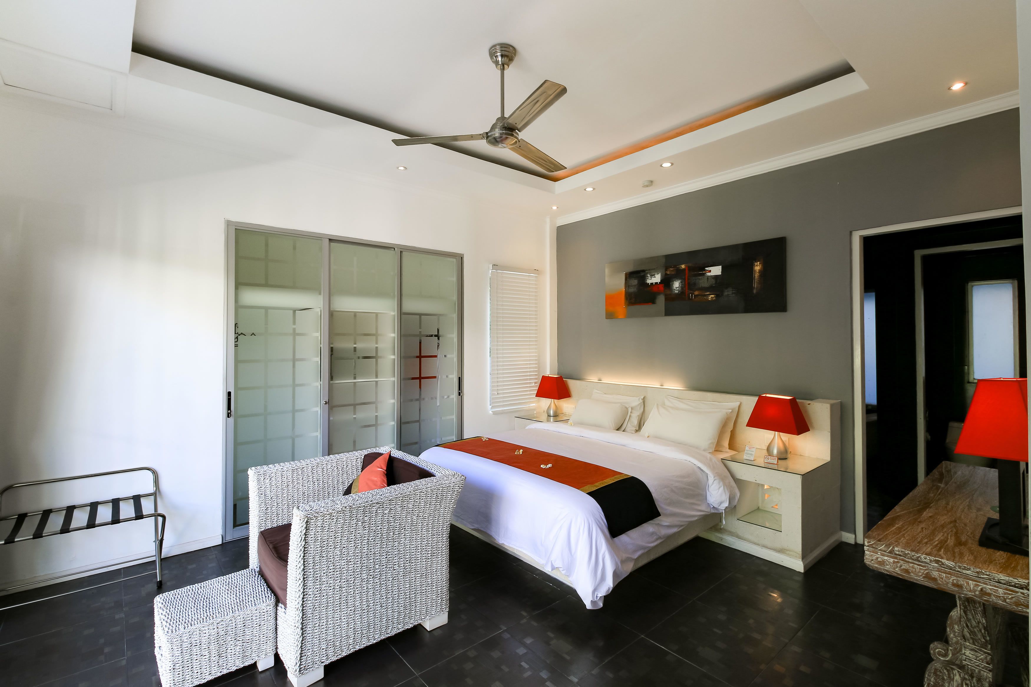 A modern hotel room with a bed, two bedside tables with lamps, a ceiling fan, a glass door leading to a bathroom, and a seating area with a chair and bench.
