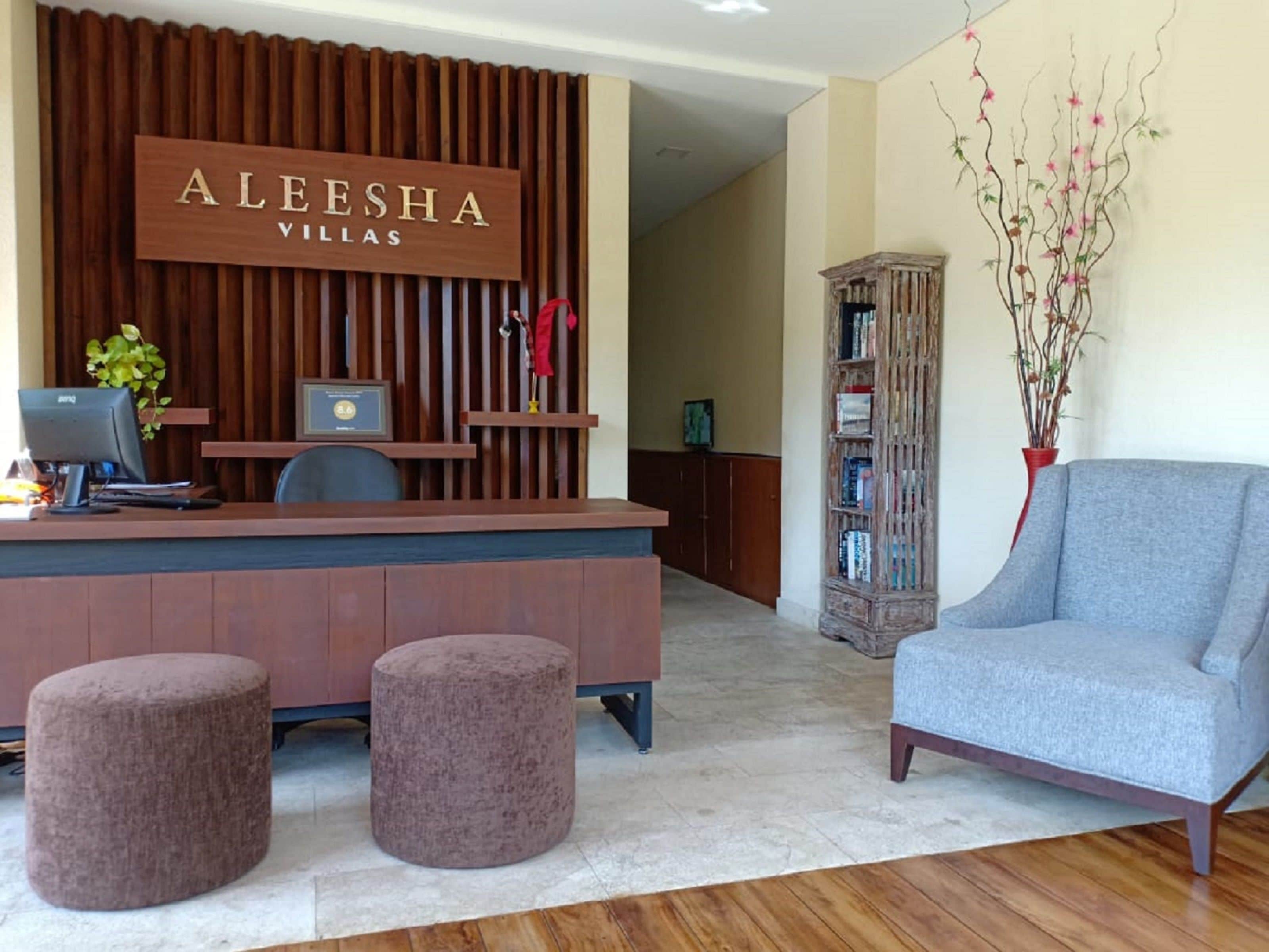 A reception area with a wooden desk, two cushioned stools, a grey armchair, a bookshelf, and a sign that reads "ALEESHA VILLAS".